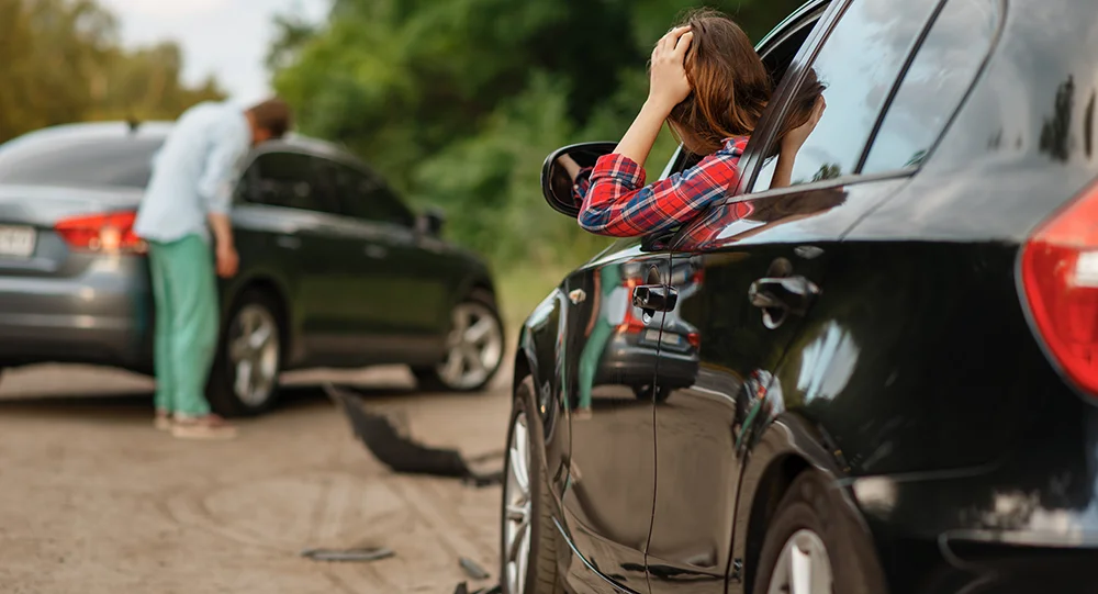 Eine Frau sitzt im Auto und hält ihren Kopf nach einem Autounfall. Im Vordergrund ist ein zweites Auto mit einem Mann, der den Schaden an seinem Fahrzeug anguckt